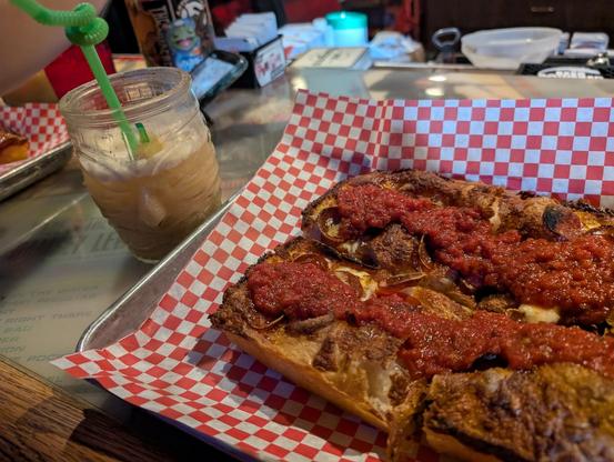 Detroit style pizza on a metal tray with red and white checkered paper, next to a glass tiki mug with a painkiller cocktail that has a green twisty straw