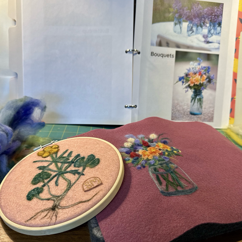 Two needle felted pieces in the foreground: botanical style buttercup on pink background and colourful bouquet in jar on plum background. Behind these is a binder holding reference photos of bouquets and a bundle of blue wool roving.