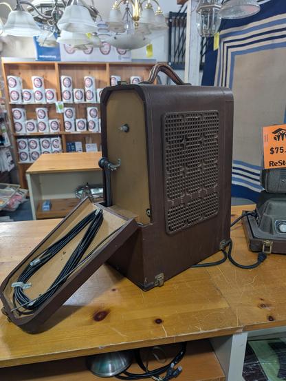 The case for the projector in the other picture, a brown wooden lid that fits over the top and latches at the bottom, with a carry handle on top. Part of one end is open with accessories inside. There is an elaborate grill on the side that covers a speaker.