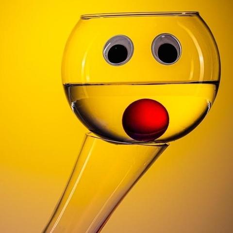 Glass bowl filled half with water and attached glass eyes and one red glass ball inside of it makes it look like a smiley clown face photographed against a yellow background. The bowl is resting on top of a carafe which is tilted in an angle, giving the figurine a neck.