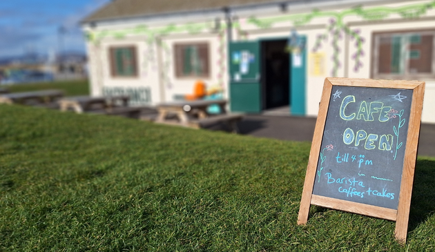 Front in focus, a green lawn and on the right a black board put up, saying that the Cafe is oprn till 4:00pm, Barista Coffees + Cakes.
In the background a one storey buildung with chairs and benches in front, an open door, walls decorated with what may be green leaves on a white-yellow ground.