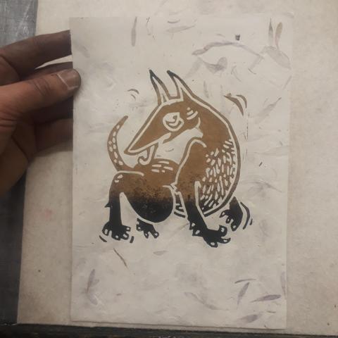 Ma grosse main pleine d'encre tient une feuille de papier incrustée de feuilles d'arbre. Dessus est imprimée une lino de chien bizarre, type naif médiéval, qui se lèche le bas du dos en gigotant. Ses pattes et le haut de ses oreilles sont noires, le reste est doré.