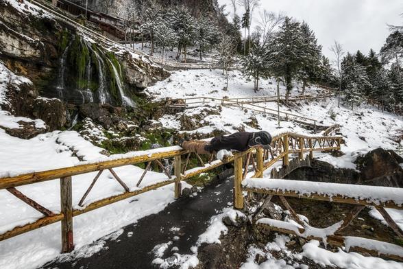 Planking über Holzgeländer in einer schneebedeckten Parklandschaft mit Wasserfall