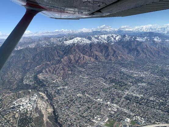 Aerial view. Below the wing of the airplane, a snow-capped mountain range with a few clouds. In the foreground, a suburban town. Close inspection reveals many of the buildings in the town are destroyed. At lower left, a dry riverbed and industrial-looking campus (it’s the Jet Propulsion Laboratory)