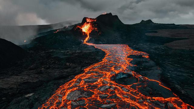 Black rock volcano with a grey smoggy sky above. Hot lava spurts out of the centre of the photo and flows towards the viewer. The lava river is red hot glowing between the cooler black crusts floating along.