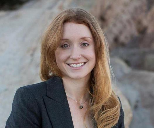 Young woman wearing a dark grey suit jacket. Medium long light brown hair flows over here left shoulder. She smiles. This is Jess Phoenix, a volcanologist.