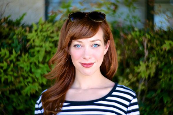 A young fresh faced woman stands in front of green bushes. She wears a black and white stripey top. She has piercing blue eyes and full red lips in a smile. She has medium long warm red-brown hair falling over her right shoulder. This is Alie Ward, creator of the podcast 'Ologies'.