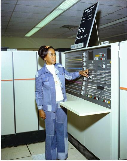 Annie Easley in a stylish blue denim outfit stands next to a large computer console, adjusting controls. The setting is an office with overhead lighting and multiple computer units.