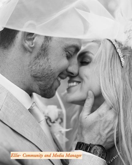 Black and white photograph of a newlywed couple sharing an intimate moment under the bride’s veil. The groom, wearing a light-colored suit and tie, gently holds the bride’s face as they lean in with their noses touching and eyes closed. The bride, wearing a sparkling tiara, smiles warmly. The groom’s wristwatch is visible on his left hand. At the bottom of the image, a text overlay in an elegant orange font reads, 'Ellie - Community and Media Manager.'