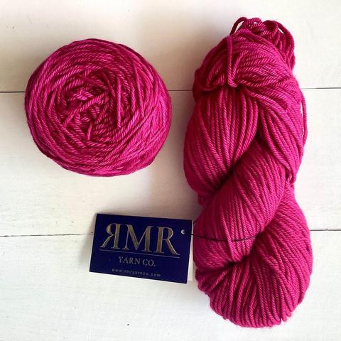 Flat lay view of dark pink yarn on a white plank background. There is one skein, and one cake of the yarn.