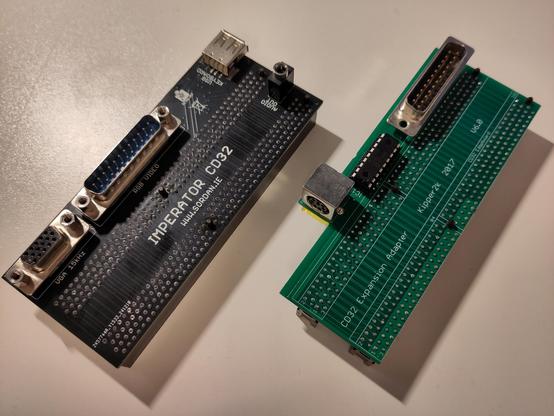 A sordan.ie "Imperator CD32" adapter and a Kipper2k CD32 adapter side by side, showing the side that faces outside the console. The sordan.ie adapter sports a DSub VGA, a 23 Pin Amiga RGB port, an USB port for the keyboard, and a 3.5mm jack audio output. The Kipper2k adapter has a PS/2 keyboard port and a 23 Pin Amiga RGB port.