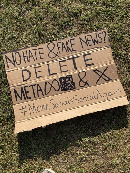 A sign laying on the grass. It’s made of cardboard and a black marker. The sign says “No hate & fake news? Delete Meta & X #MakeSocialsSocialAgain” and it has the symbols of Meta, Facebook, Instagram, WhatsApp, Threads and X on it.