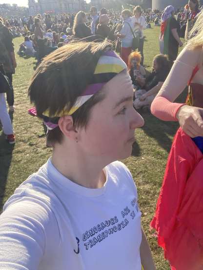 A photo of me at the Feminist March. I’m making a selfie, while looking away from the camera. I’m wearing a bandana in the colors of the nonbinair flag: yellow, white, purple and black. And a white t-shirt with the text “Dinosaurs are cool. Transphobia is not.” on it with a small dinosaur next to the text. I’m standing on a field, filled with people in the background.
