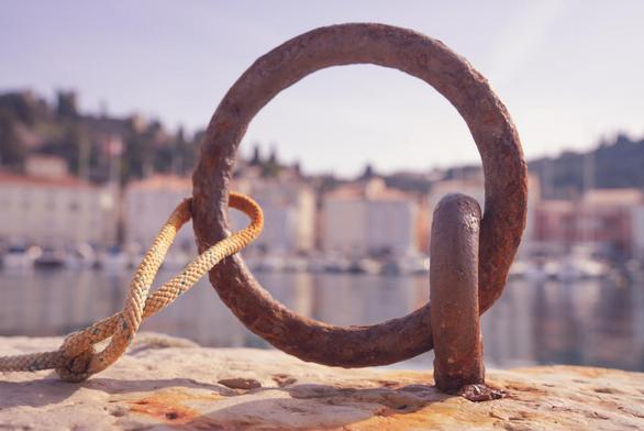 Ein Mooringring am Hafen von Piran, der Ring ist sehr rostig und steht hochkant, so das man durchsehen kann. Links sieht man das ein Stück einer Leine, im Hintergrund der Hafen und die Gebäude von Piran.