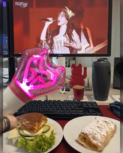 photo of an office desk with some breakfast in front of the keyboard, a taeyeon concert on the computer screen and a hand holding a kpop cheering lightstick.