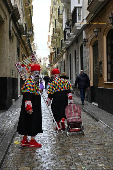 imagen en formato vertical donde aparecen dos hombres vestidos para el carnaval, con birretes rojos y encajes en las bocamangas y mucetas de rombos multicolores.  Uno de ellos camina de espaldas a la cámara por la mitad de la calle arrastrando un carrito de la compra. El otro, se ha detenido un momento para mirar hacia atrás. Porta un pequeño cartel donde puede leerse «Los catedráticos del carnaval»
