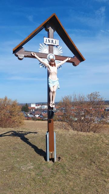 Ein Kreuz mit Christusfigur und Aufschrift "I.N.R.I." auf einer Anhöhe über einem Ort. Ein sonniger Tag.