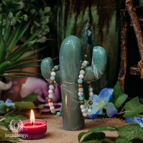 Two examples of the exclusive Balance Gemstone Bracelet hang from a carved green aventurine saguaro cactus with a burning votive candle for scale. The bracelets are elastic and have no clasp. They consist of small round spherical beads of Amazonite, Unakite, and Howlite. This is an exclusive product from Inked Goddess Creations.