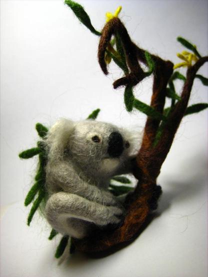 Brooch made of nettle fibres by dry felting, in the form of a koala. The koala sits on a branch of a blossoming eucalyptus and holds onto it with one front and one back paw. It is resting.