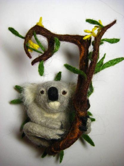 Brooch made of nettle fibres by dry felting, in the form of a koala. The koala sits on a branch of a blossoming eucalyptus and holds onto it with one front and one back paw. It is resting.