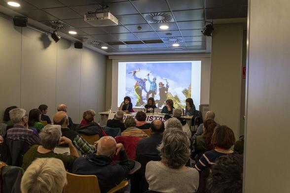 Sala llena con aproximadamente 70 personas ante la mesa de 4 ponentes y al fondo una imagen de tres mujeres montañeras
