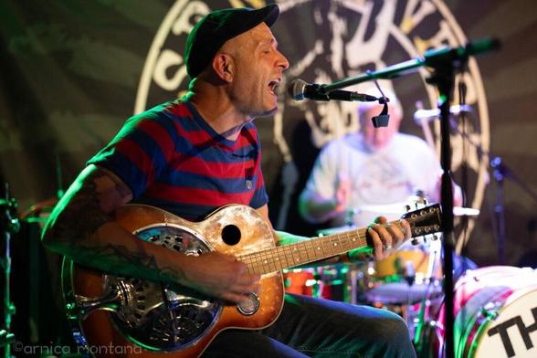 A musician wearing a striped shirt and hat performs on stage, singing passionately into a microphone while playing a resonator guitar. In the background, a drummer is partially visible, contributing to the live music atmosphere.