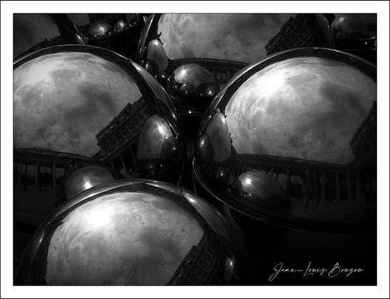 The image features a group of polished metallic spheres that create a striking visual display. Their reflective surfaces create a mesmerizing interplay of light and shadow, reflecting the surrounding environment in a distorted yet intriguing way. You can observe architectural details and perhaps clouds mirrored in the orbs, which introduces a sense of depth and invites contemplation.
The monochromatic palette adds a layer of sophistication and drama, stripping away distractions and allowing the viewer to focus on the shapes and the reflections. This choice might evoke feelings of nostalgia or contemplation, as it emphasizes contrasts between light and dark, smoothness and the implied texture of their surroundings.
