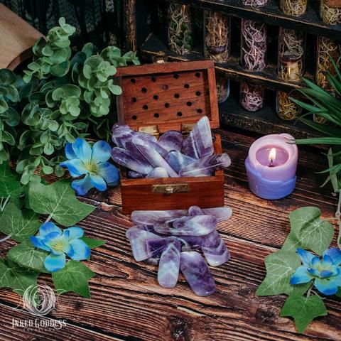 A handful of nuggets of the "Banded Amethyst Gemstone Slice" product are arranged in front of a small wooden box holding several more specimens. They are polished smooth and in shaded bands of purple and white. Presented by Inked Goddess Creations.