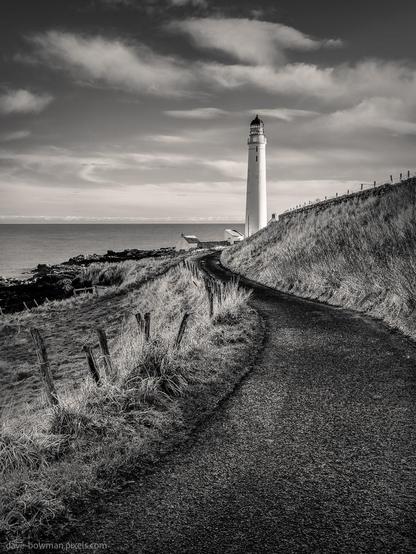 A photograph of the road leading to Scurdie Ness lighthouse in Montrose, Angus, Scotland. The image emphasises the bold contrast and textures of the landscape, with the long, empty road guiding the viewer’s gaze toward the lighthouse, which stands boldly at the horizon. The monochrome tones accentuate the simplicity and strength of the scene.