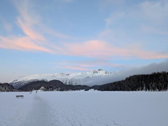 Lac de St Moritz gelé recouvert de neige avec un sentier piéton. Ciel grandiose avec quelques nuages rosés dans le coucher du soleil et des sommets enneigés