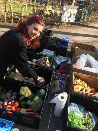 Anika Liekefett beugt sich über Kisten, die mit Gemüse gefüllt und auf einer Tischtennisplatte aufgestellt sind. Sie reinigt den Boden einer Kiste, um dort gleich etwas hinein zu sortieren. Außerdem lächelt sie freundlich in die Kamera. In einer anderen Kiste liegen Tomaten, Brokkoli und rote Bette, in einem Karton sieht man Bananen. In der Mitte steht eine Küchenrolle.

Wenn wir Sonnenschein Wetter haben, dann machen wir an Donnerstagen, passend zu unserem Logo, einer innig lächelnden und freundlich strahlenden Sonne, die Verteilung geretteter Lebensmittel draußen, vor dem HausDrei Eingang.

Wir bauen Sitz-Bänke auf und nutzen die Tischtennis-Platte vor dem Kindertreff Spielplatz, um dort die Kisten und Kartons aufzustellen.
Wir machen Heißwasser fertig und haben Tee-Beutel mit. Aus der Küche leihen wir uns Tassen. Wenn in den geretteten Waren Kuchen ist, teilen wir den gerne sofort auf und schon wird aus der Lebensmittelverteilung eine gemütliche, gesellige Teatime, wie es im Angelsächsischen heißt.

Anders als an den anderen Tagen, haben wir bei „Retten Teilen Helfen“ an Donnerstagen sehr viele gerettete Lebensmittel aus Bio-Märkten. Aber auch Sachen vom Supermarkt. Selten ist etwas vom Vortag, vom Discounter übrig. Und wenn, dann nur so etwas, wie der sehr beliebte Sellerie oder farbenreiche, reife Bananen.
