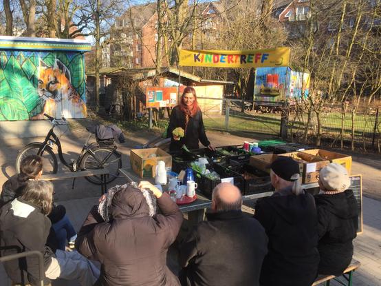 Anika Liekefett steht hinter einer Tischtennisplatte und hält einen Brokkoli hoch. Auf der Platte stehen mehrere Kunststoffkisten und Pappkartons, die mit geretteten Lebensmitteln gefüllt sind. Davor sitzen einige Gäste auf Sitzbänken. 

Bei „Retten Teilen Helfen“ haben wir von den verschiedenen Sorten nicht oft große Mengen. Die wenigen Brokkoli, Karotten, rote Beeten, Zucchini, Pilze, Pastinaken, Gurken, Fenchel, Chicorée, Blumenkohl, Zwiebeln, Kartoffeln, Süßkartoffeln, Weißkohl, Tomaten, Salate aller Art, Äpfel, Pampelmusen, Melonen, Erdbeeren, Himbeeren, Blaubeeren oder Apfelsinen müssen friedlich und gerecht unter allen Gästen verteilt werden.

Um Gerangel zu vermeiden, haben wir uns deshalb gemeinsam dafür entschieden, in einer Reihenfolge aus den Kisten Waren zu entnehmen. Nacheinander bekommt jede*r ein oder zwei Teile die er/sie aussucht. Wenn alle einmal durch sind und noch etwas übrig ist, geht es wieder von vorne los. Wer später kommt wird ans Ende der Reihe gesetzt.

Anika Liekefett und ein*e Helfer*in moderieren das Ganze. Teilweise sind Backwaren dabei, die sollen nur mit Einmal-Handschuhen berührt werden.
Für Verteilungen bei „Retten Teilen Helfen“ sind keine An- oder Abmeldungen nötig. Wer kommt, kommt, wer geht, geht. Das Einzige, was mitgebracht werden sollte, ist Frieden, Freundlichkeit, Respekt und kooperatives Miteinander. Wir machen alles mit einer zarten, sanften Geschwindigkeit, ohne Hast, ohne Termindruck.