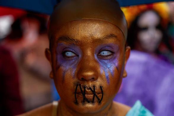 A woman with golden brown skin, azure contact lenses, blue tear streaks and "sown up" lips.