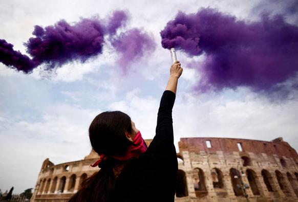 A young woman with a red scarf covering her face  holds a purple smoke flare in front of the Colosseum.