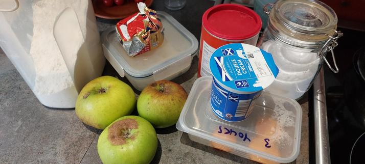 On a grey kitchen worktop next to a cooker is the following: a jar of caster sugar, 3 egg yolks in a plastic tub labeled "3 yolks", a pot of double cream on top of the eggs, a tub of cornflour, three cooking apples, a large plastic container of flour, and a glass container with a small amount of crumble topping in it and a thing of butter on top. Making custard and apple crumble for later.