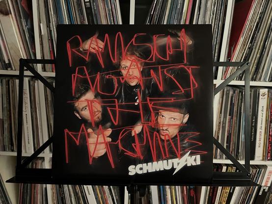 Ein Schallplattencover mit dem Bandnamen "SCHMUTZKI" und dem Titel "Ramsch Against the Machine", geschrieben in fettem rotem Text. Das Bild zeigt drei männliche Bandmitglieder, teilweise verdeckt durch die Graffiti-ähnliche Schrift.
