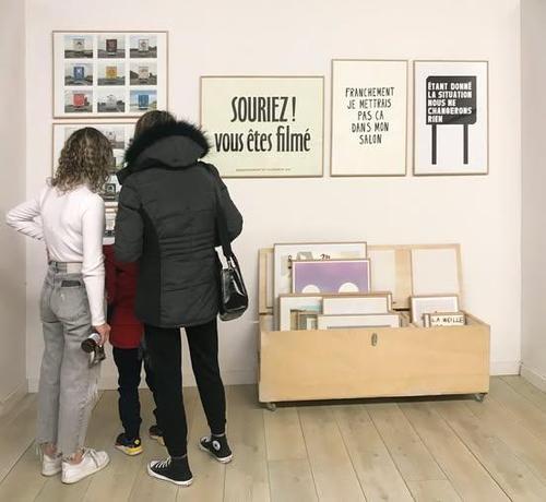 Trois personnes observent des œuvres d'art accrochées au mur. Un bac au sol contient d'autres œuvres encadrées au sol.
