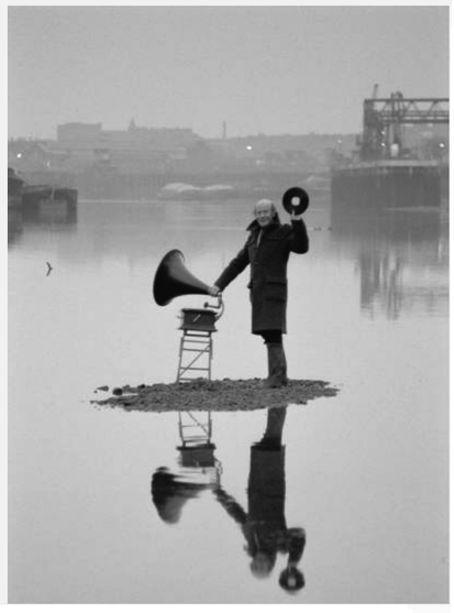 Man on a small island in a body of water, holding a gramophone and a record, with a reflection in the water.