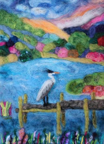 A vibrant needle-felted artwork showing a heron standing on a wooden pier over a blue lake. The scene is surrounded by lush green hills, colorful trees and distant mountains with warm hues in the sky. The foreground features bright reeds and reflections in the water giving the piece a textured, dreamy feel.