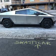 Photo of a Cybertruck Tesla car parked in New York. Someone wrote 'Swasticar" with an arrow, pointing in the direction of the car, on the street with sidewalk chalk.