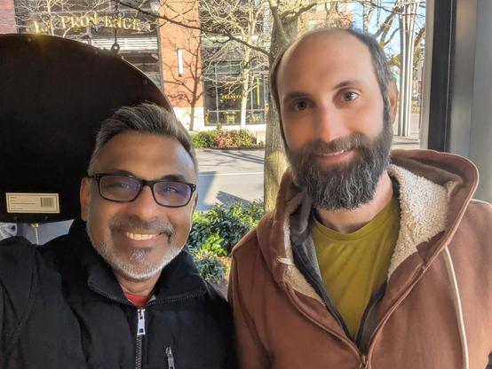 An indian man with white and black hair standing next to a taller white gentleman with a beard and moustache. Both are smiling.