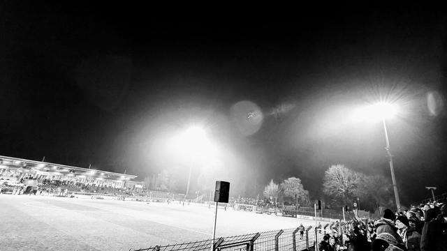 Schwarz-Weiß-Foto von den Rängen eines Stadions auf das Spielfeld. Der Himmel ist dunkel, das Spielfeld wird von Flutlicht erhellt. Zwei Flutlichtmasten sind vor dem dunklen Himmel zu sehen.

Black and white photograph taken from the stands of a stadium and looking onto the pitch. The sky is dark and the pitch is lit by floodlights. Two floodlight columns can be seen against the dark sky.