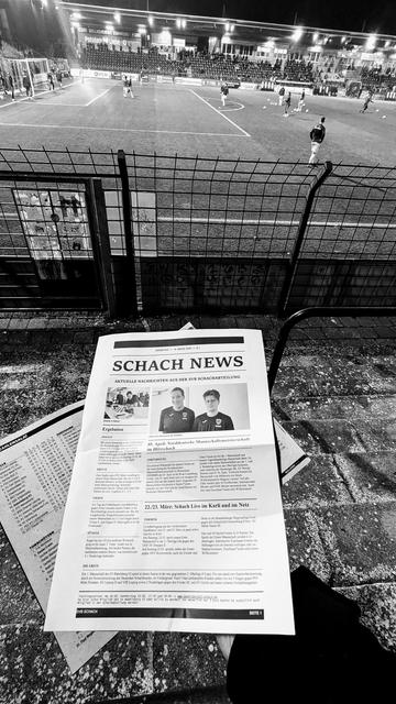 Schwarz-Weiß-Foto von den Rängen eines Stadions auf das Spielfeld. Im Vordergrund hält eine Hand ein Heft. Auf dem Heft liegt ein Zettel auf dem Artikel und Fotots zu sehen sind. Die Überschrift auf dem Zettel lautet: "Schach News"

Black and white photograph of the stands of a stadium looking onto the pitch. In the foreground, a hand is holding a magazine. On the magazine, there is a piece of paper with articles and photos. The title on the paper reads: ‘Schach News’