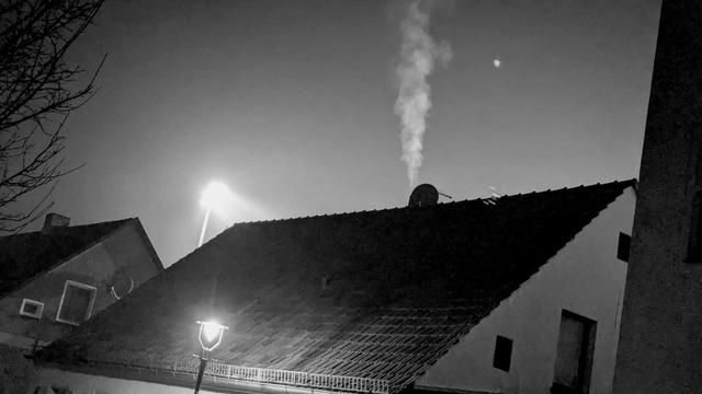 Schwarz-Weiß-Foto. Vor einem dunklen Himmel sieht man im Vordergrund die Dächer von Häusern. Aus einem Kamin steigt Rauch senkrecht in den Himmel. Erleuchtet wird die Szene von einem Flutlichmast der im Huintergrund zu sehen ist.

Black and white photograph. Against a dark sky, the roofs of houses can be seen in the foreground. Smoke rises vertically from a chimney into the sky. The scene is illuminated by a floodlight mast that can be seen in the background.