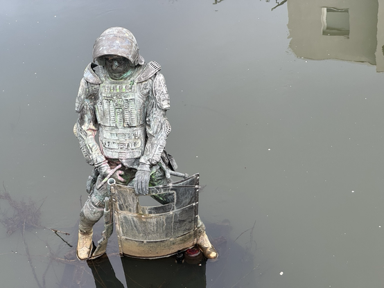 Standbild, dass einen Mann in Polizeirüstung darstellt. Das Standbild steht mitten im Flüsschen Hase und scheint urinieren zu wollen. Im langsam fließenden Wasser spiegelt sich ein Stadthaus.