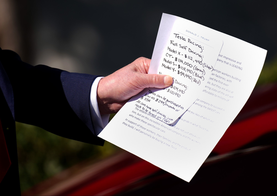 Close up photo of Trumps hand holding a note and a smaller note on top of it, with Tesla prices and details written in his own handwriting, large letters because otherwise he can't read it.