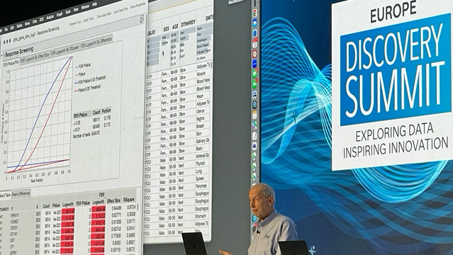Man talking on stage. On background graphs, numbers, and the blue-white logo of the JMP discovery summit.