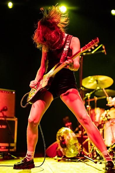 A musician with long hair is passionately playing an electric guitar on stage, illuminated by vibrant yellow lights. The performer is dressed in a black outfit and is caught mid-movement, showcasing a dynamic pose. In the background, a drum set and amplifiers.