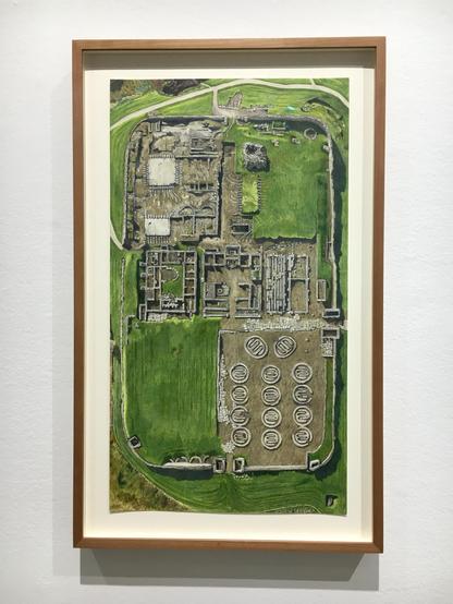 A framed drawing of a substantial building complex viewed from above and surrounded by a vibrant green field