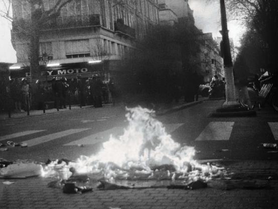 Photographie argentique en noir et blanc capturant une scène de rue chaotique : un feu brûle au milieu de la chaussée, entouré de déchets calcinés. En arrière-plan, une foule se rassemble devant un magasin, tandis que d'autres silhouettes se déplacent sur le trottoir. Une scène de tension née d’une marche féministe pacifique perturbée par des violences d’extrême droite.